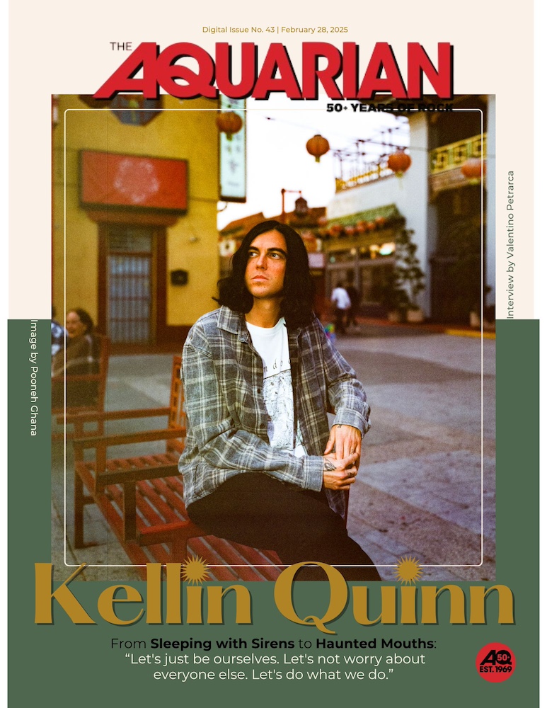 A young man wearing a flannel shirt over a white t-shirt and black jeans sittings with his legs crossed on a wooden bench. There are paper lanterns hung up on the street behind him and shops with red and green awnings. The man has long black hair and thick eyebrows. He looks to the left thoughtfully and his name is written largely in gold underneath, "Kellin Quinn." The letter 'I' in the first and last name are 'dotted' with sunbursts. The magazine's name is up top. "The Aquarian," in red.