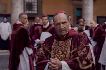A priest looks questioningly to her right while wearing his Catholic robe and yarmulke. The priests outfits are red and maroon, while other members of the clergy walk around him in white robes.