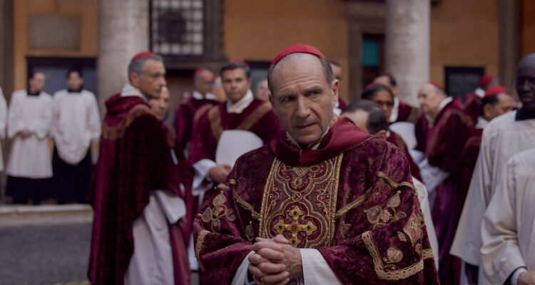 A priest looks questioningly to her right while wearing his Catholic robe and yarmulke. The priests outfits are red and maroon, while other members of the clergy walk around him in white robes.
