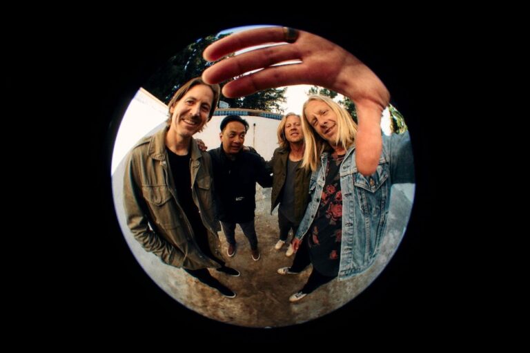 Four men stand in an empty pool and look into a camera with a round fisheye lens. The man on the right has long blonde hair and looks to be reaching for the camera, his hand just cupping the top of the frame. He is wearing a blue denim jacket. The three men next to him stand together smiling at the camera.