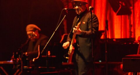 A man in a suit and playing a guitar standing on stage in front of a microphone. There are red lights lighting the stage. The man is wearing glasses and a circular brimmed hat. Behind him are lights, amps, and microphone stands. In the left corner is a man on keyboards just out of focus.