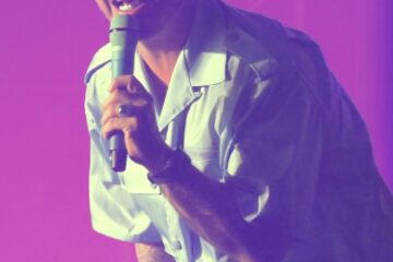 Man with bleached buzzcut hair sings into a microphone. He leans into the crowd and wears a blue button-up t-shirt. The lights around his are pink.