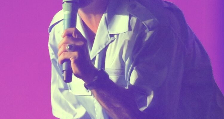 Man with bleached buzzcut hair sings into a microphone. He leans into the crowd and wears a blue button-up t-shirt. The lights around his are pink.