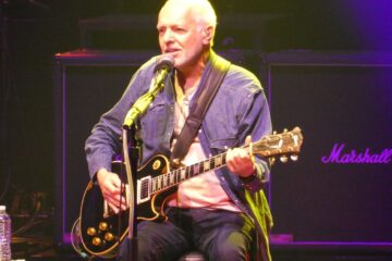 A man sits behind a microphone, singing into it and playing a guitar that is on his lap. He is older with white hair and is wearing a blue button-up shirt open with a white t-shirt underneath. He is on a stage.
