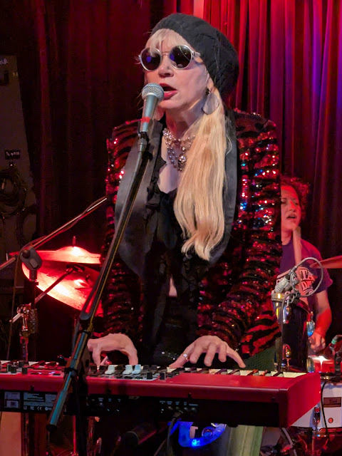 A female singer with long blonde hair under a grey slouching hat is singing at a red keyboard. She has sunglasses on and a sparkly red and black striped blazer on.