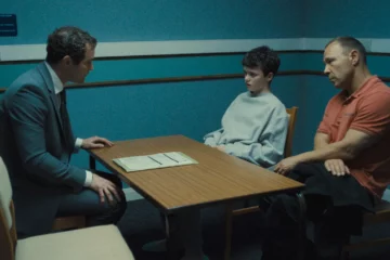 A young boy is sitting next to his dad on one side of a table. On the other side is another man, in a suit, and he seems to be questing the young boy. They are in a dimly lit office painted a dark blue.