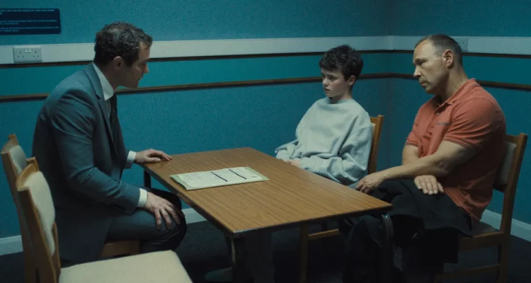 A young boy is sitting next to his dad on one side of a table. On the other side is another man, in a suit, and he seems to be questing the young boy. They are in a dimly lit office painted a dark blue.