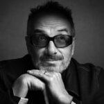 A black and white photo of a man in tinted glasses resting his chin on his hands. He is facing forward, but his eyes are looking up. He has some facial hair and is wearing a dark shirt.