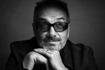 A black and white photo of a man in tinted glasses resting his chin on his hands. He is facing forward, but his eyes are looking up. He has some facial hair and is wearing a dark shirt.