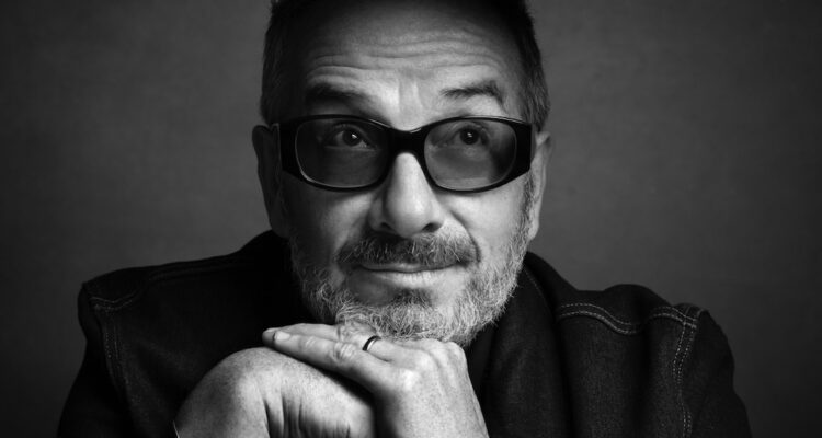 A black and white photo of a man in tinted glasses resting his chin on his hands. He is facing forward, but his eyes are looking up. He has some facial hair and is wearing a dark shirt.