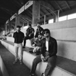 Fix men sit on concrete bleachers. It is a film photo with cracked/leaked edges and is in black-and-white. They all have facial hair, and the man closest to the camera wears sunglasses.