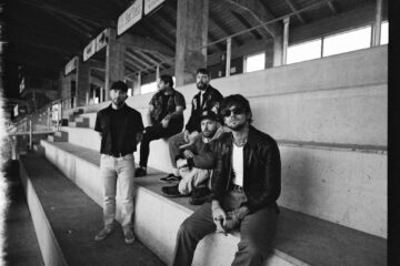 Fix men sit on concrete bleachers. It is a film photo with cracked/leaked edges and is in black-and-white. They all have facial hair, and the man closest to the camera wears sunglasses.