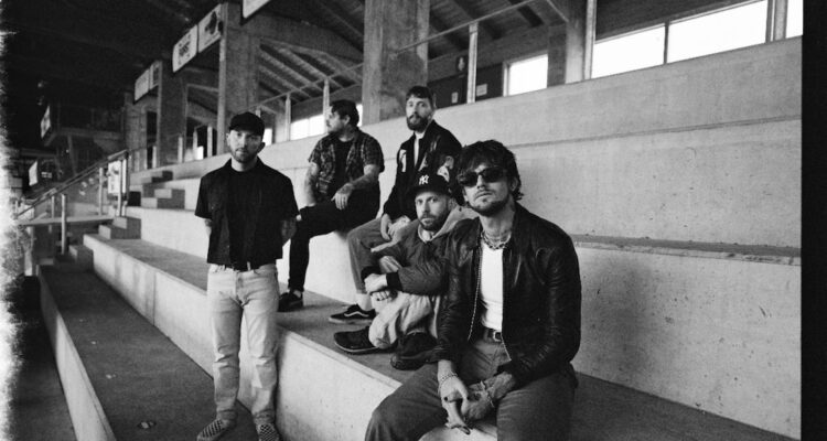 Fix men sit on concrete bleachers. It is a film photo with cracked/leaked edges and is in black-and-white. They all have facial hair, and the man closest to the camera wears sunglasses.