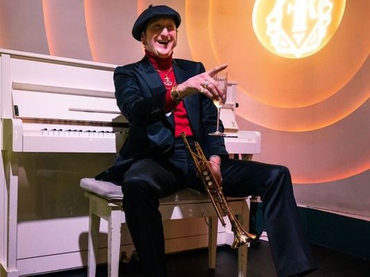 A man smiles and points to the right, wearing a red button-up shirt and a black suit with a matching black beret on his head. He is holding a trumpet and sitting at a white grand piano.